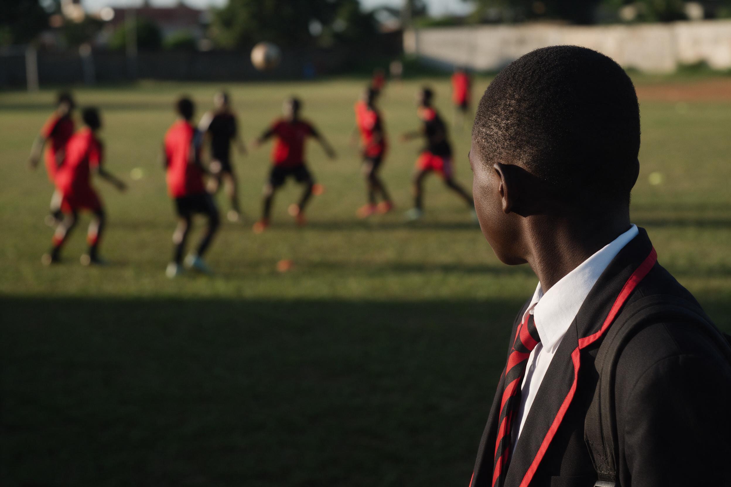 Image générée provisoire : un élève en uniforme scolaire regarde, de dos, un entraînement de football en arrière-plan.